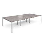Large modular bench desk with a grey wood-effect top, integrated cable access slots, and silver metal legs, shown on a white background.