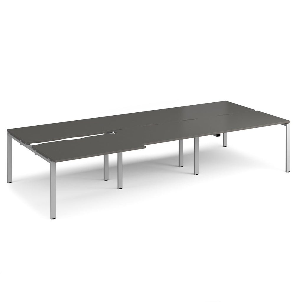 Large modular bench desk with a dark grey worktop, integrated cable access cut-outs, and silver metal legs, shown on a white background.
