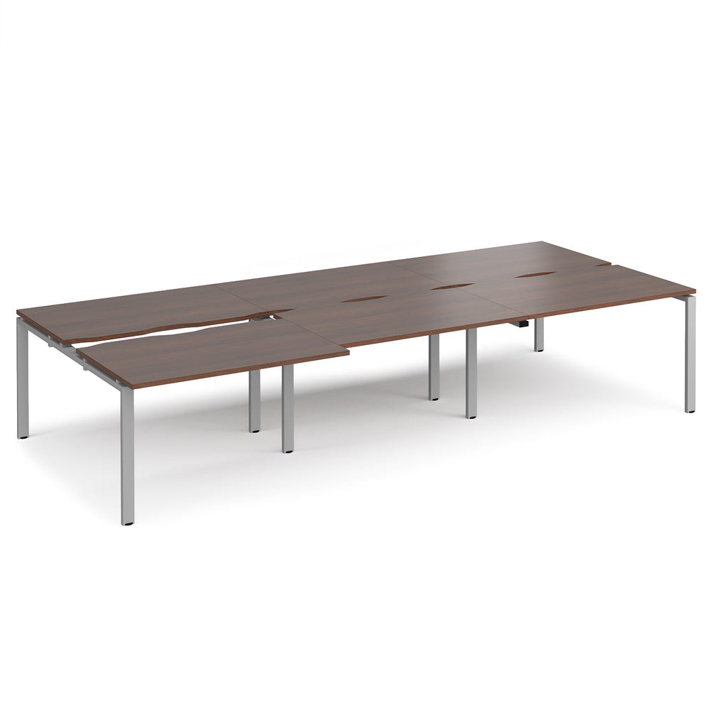 Large modular bench desk with a dark wood-effect top, integrated cable access cut-outs, and silver metal legs, shown on a white background.