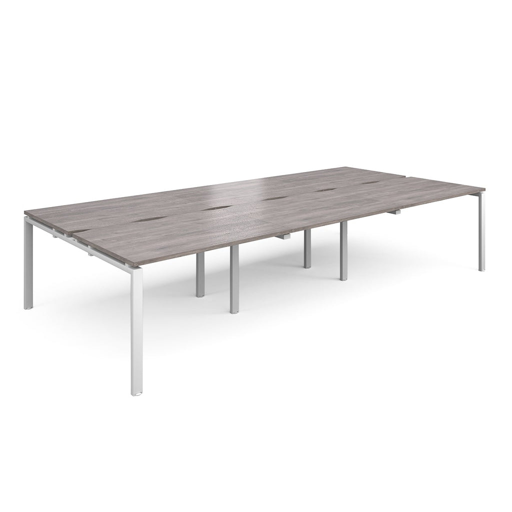 Large modular bench desk with a grey wood-effect top, integrated cable access slots, and white metal legs, shown on a white background.