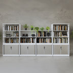 Row of white office bookcases with open shelving and lockable tambour doors, filled with books and decorative plants, arranged against a concrete-style wall.