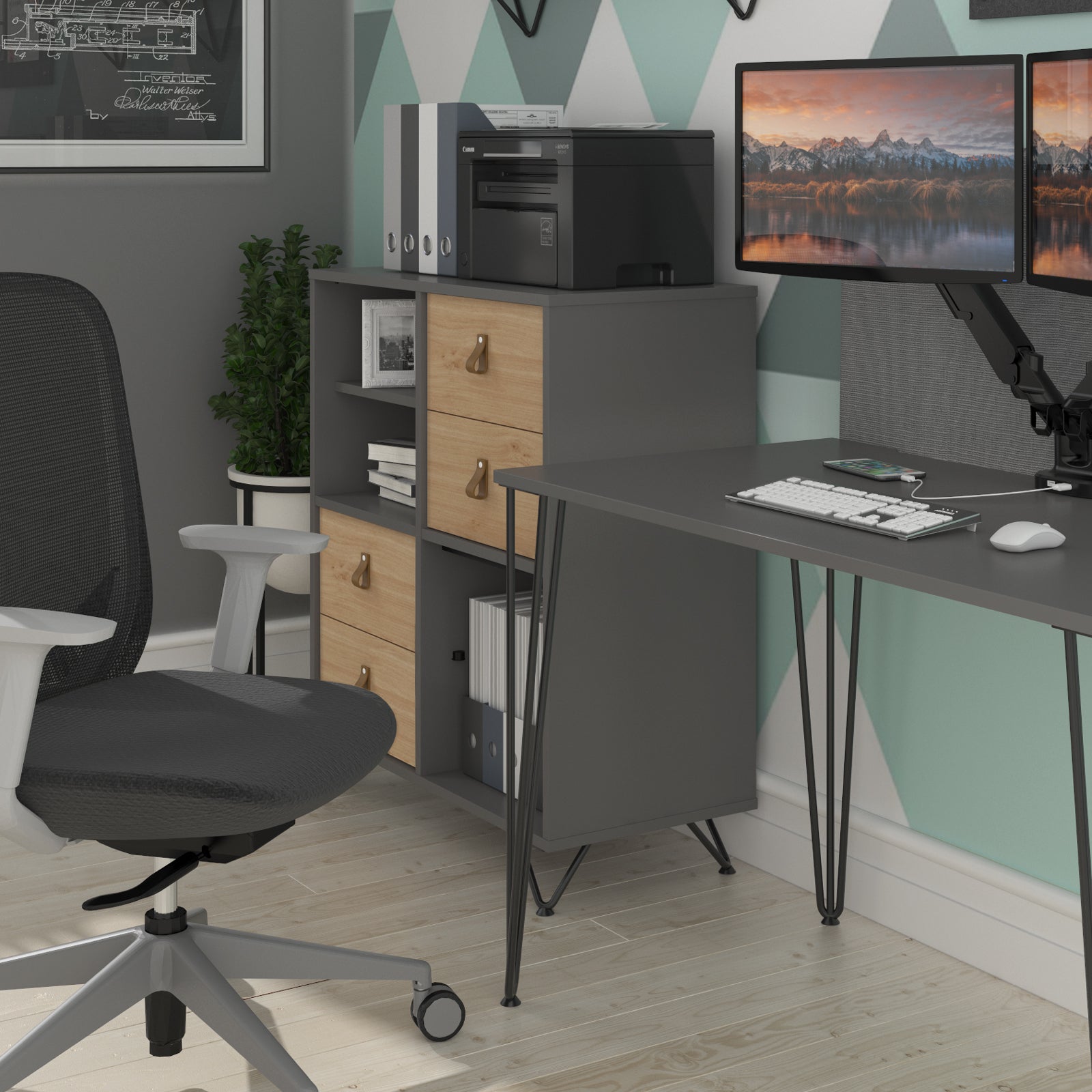 Modern home office corner featuring a grey desk with hairpin legs, matching storage cabinet with wood-effect drawers, ergonomic office chair, and dual monitors against a geometric accent wall.