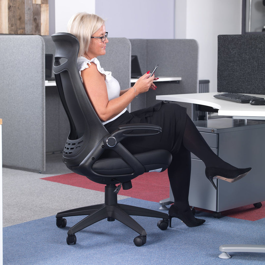 Woman seated in a black ergonomic office chair at a modern workstation, using a smartphone, with desk screens and acoustic panels in an open-plan office setting.