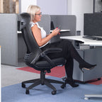 Woman seated in a black ergonomic office chair at a modern workstation, using a smartphone, with desk screens and acoustic panels in an open-plan office setting.
