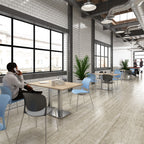 A modern office or café interior with light wood flooring, white brick walls, large black-framed windows, and multiple dining tables with blue, grey, and black chairs. A man sits at a table near the left window, and other people are visible in the background.