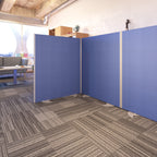 Free-standing blue office screen panels arranged in a corner configuration with metal feet, used to create a flexible partitioned workspace on a carpeted office floor.