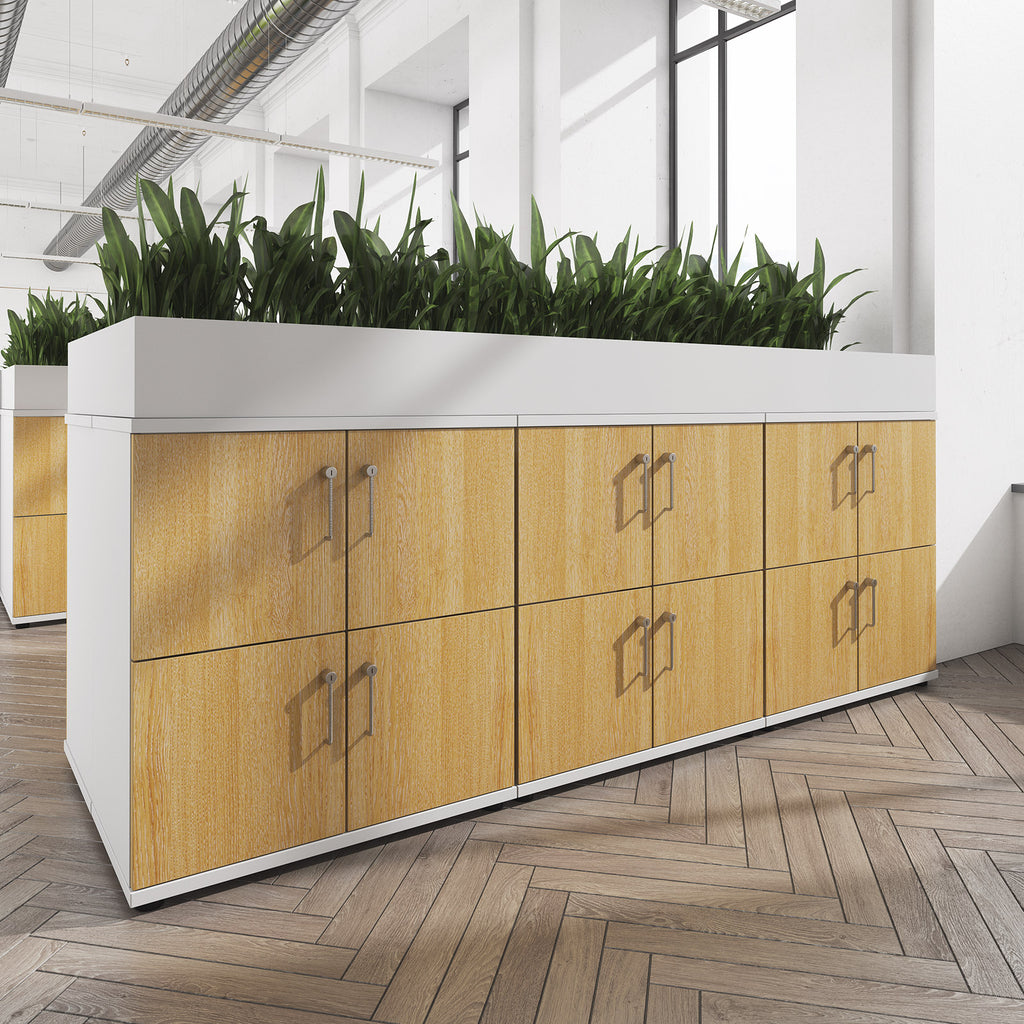 Office storage cabinet with wood-effect doors and metal handles, topped with an integrated white planter filled with green plants, set in a modern open-plan workspace.