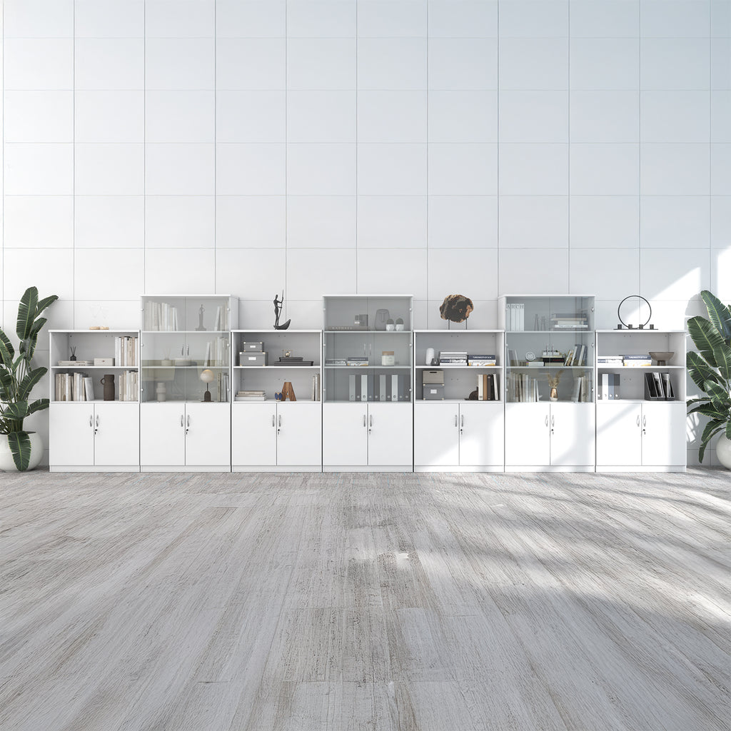A long modular white storage cabinet and shelving unit set against a white paneled wall, with light wood flooring and potted plants on both sides.